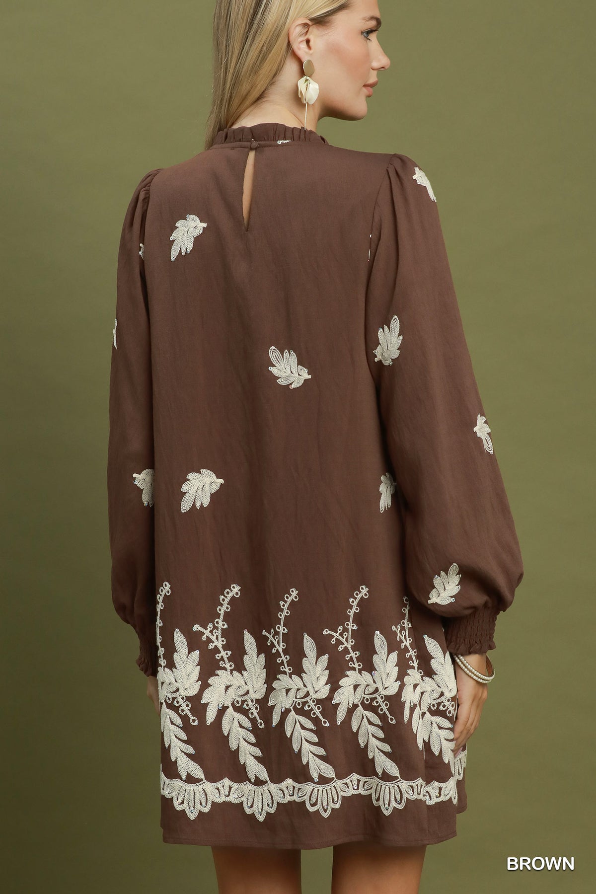 A woman shown from the back wearing a brown long-sleeve dress with white floral embroidery and a lace hem, set against a solid olive green background.