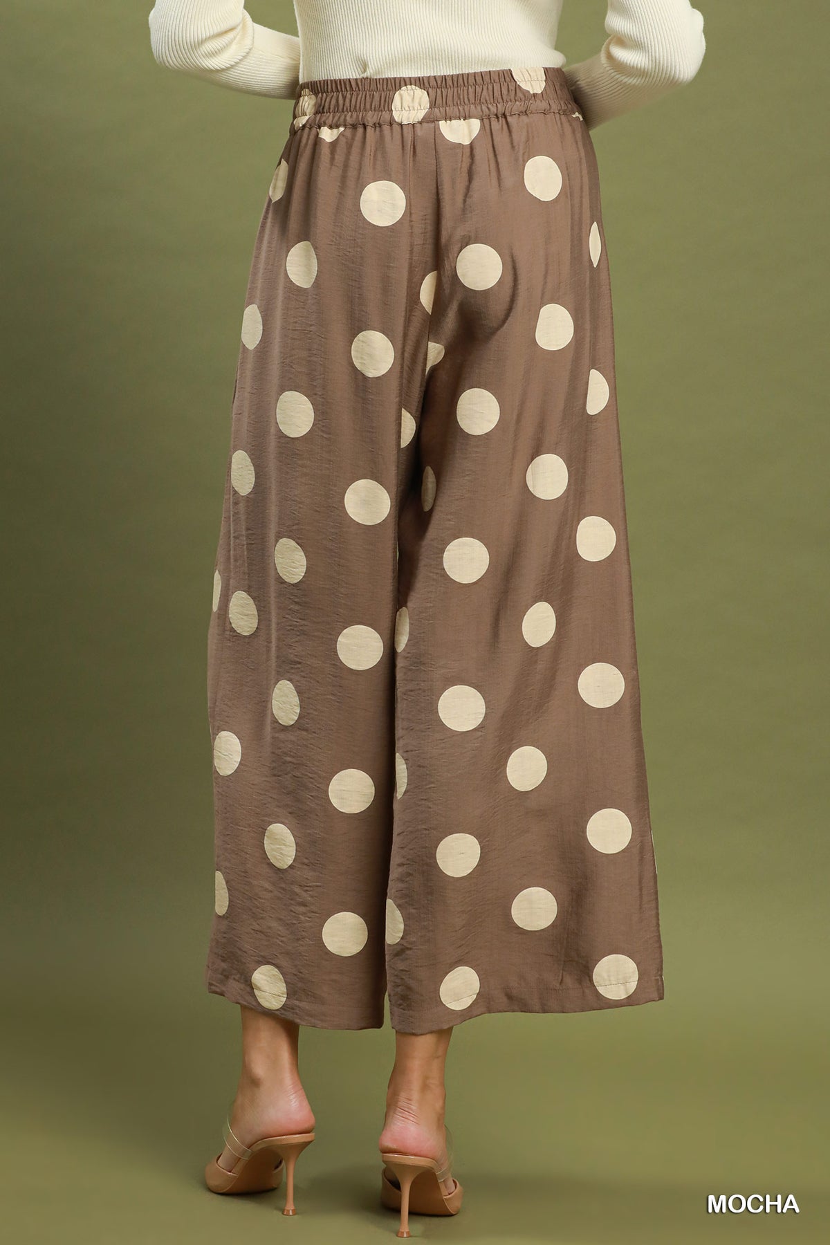 A person shown from the back wearing wide-leg cropped pants in a warm taupe-brown color with large cream polka dots, paired with a white long-sleeve top and nude heels, against an olive green studio background.